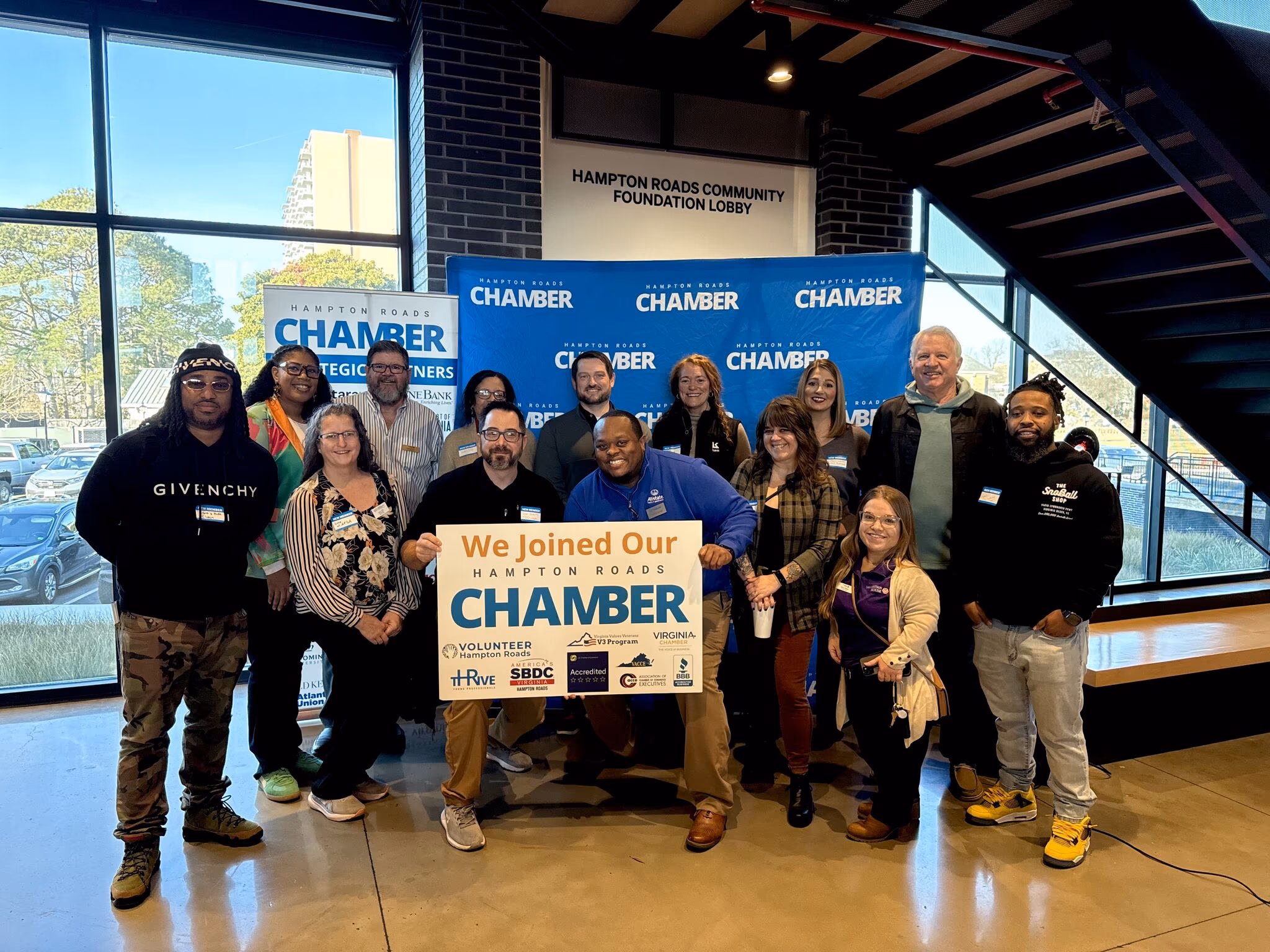 Group of diverse individuals celebrating their membership in the virginia Chamber, holding a sign that reads "We Joined Our virginia Chamber," with a backdrop featuring the Chamber's branding and community engagement themes.